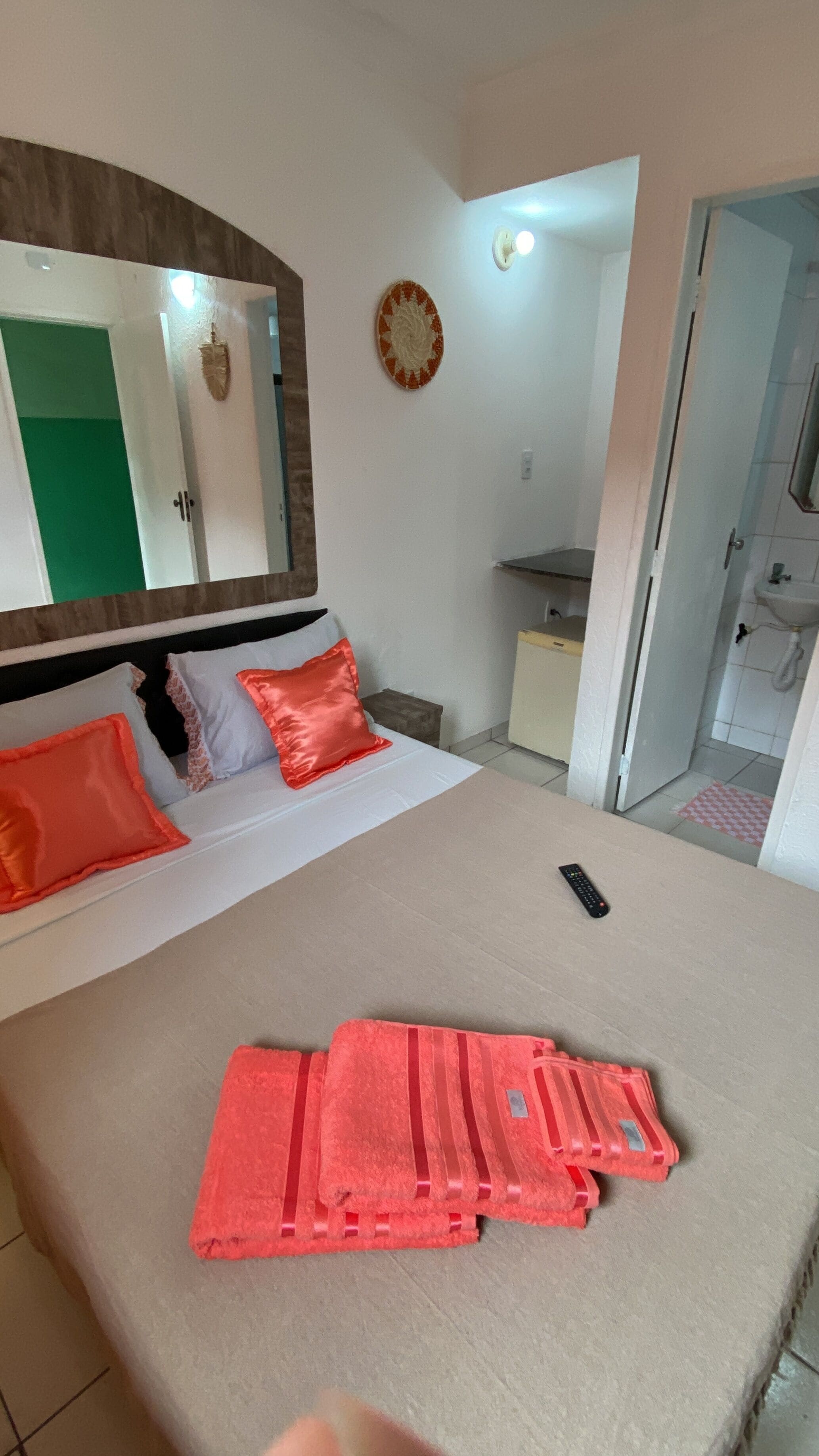 A bedroom with a bed, pillows, a bedside table, a mirror, a wall decoration, and a bathroom visible through an open door.