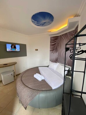 A modern bedroom with a round bed, a television mounted on the wall, a small desk, and a ceiling mural. - SSA33 Hotel (Salvador)