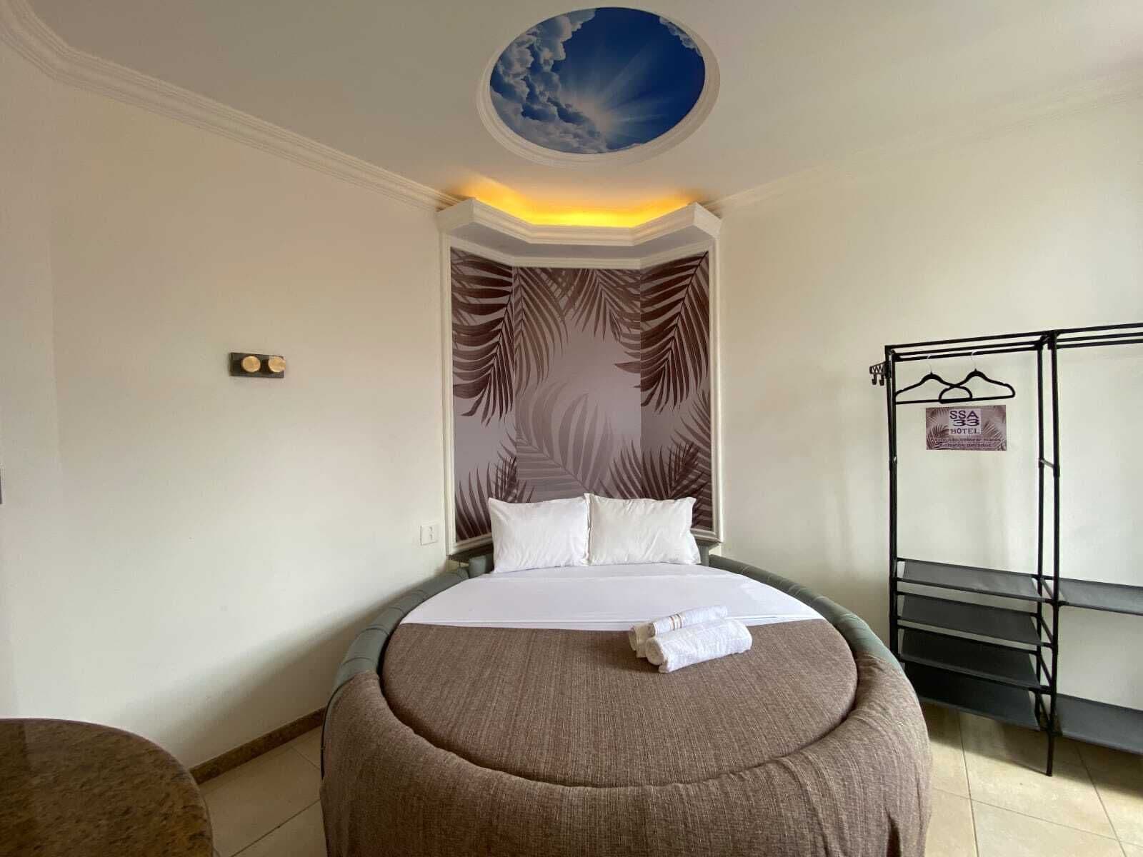 A hotel room with a bed, a round ottoman, a wall with a leaf pattern, a ceiling with a cloud design, and a rack with hangers.