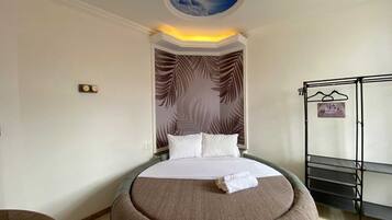 A hotel room with a bed, a round ottoman, a wall with a leaf pattern, a ceiling with a cloud design, and a rack with hangers.