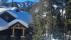 Eine schneebedeckte Holzhütte, umgeben von Bäumen und Bergen im Hintergrund.