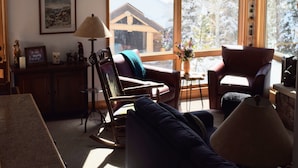 Ein gemütliches Wohnzimmer mit Holzboden, einer Couch, einem Sessel, einem Tisch mit Lampe und einem Fenster mit Blick auf ein Haus und Bäume.