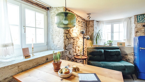 Een knus interieur met een houten tafel, een groene bank, een stenen muur en een blauwe koelkast.