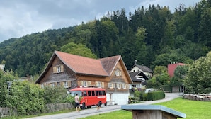 Een houten waterpomp in een grasrijke omgeving, met een rode bus op de achtergrond en huizen met rode daken.