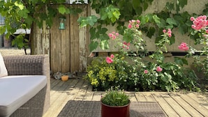 Een houten terras met rieten meubilair, een bloempot en een tuin vol groene bladeren en roze bloemen.