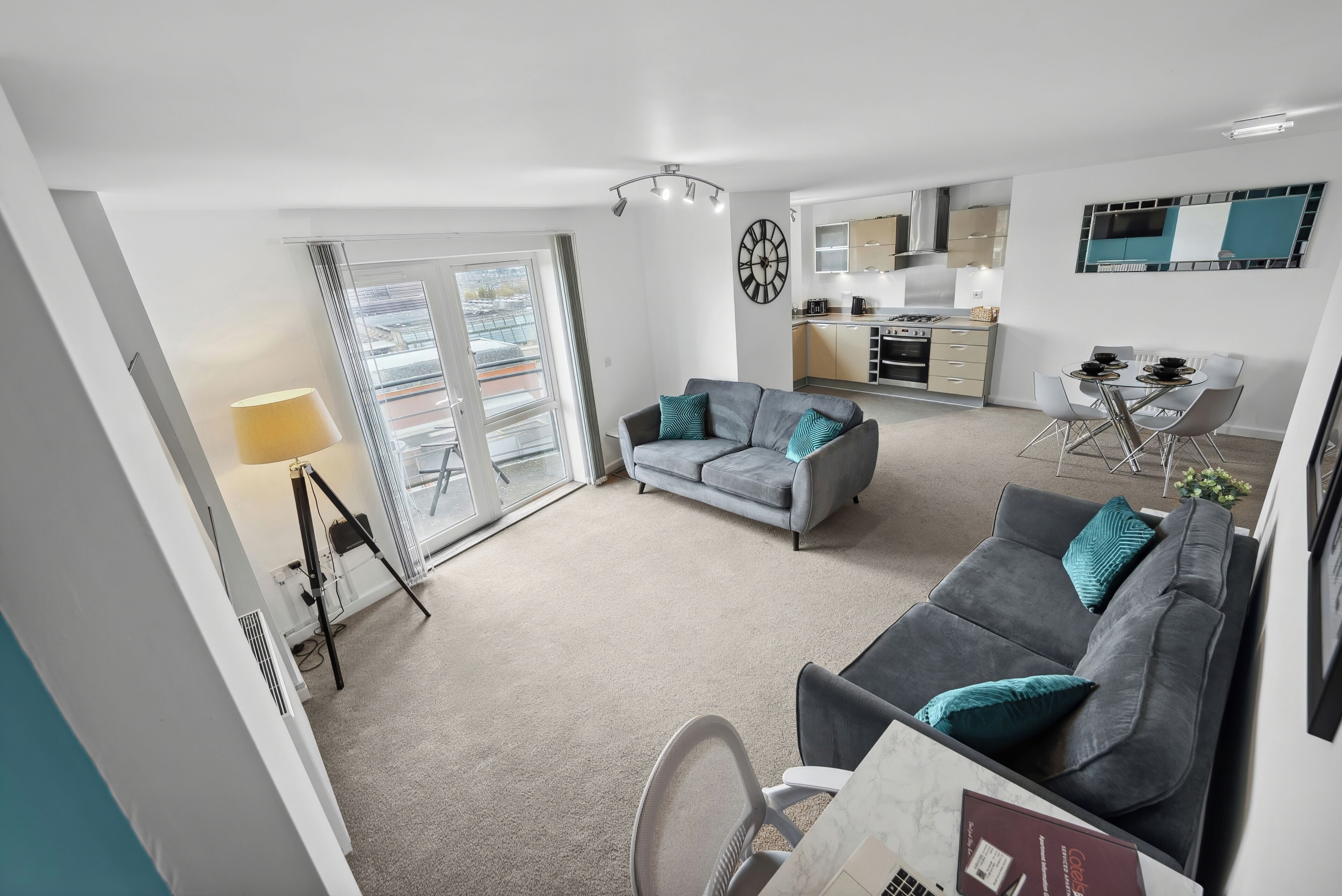 A modern apartment with a grey sofa, a dining table with chairs, a kitchen area, and a large window with blinds.
