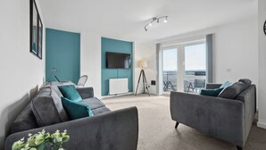 A modern living room with grey sofas, a flat-screen TV, a tripod lamp, and a view of the outdoors. - The Pinnacle by Cotels (Northampton)