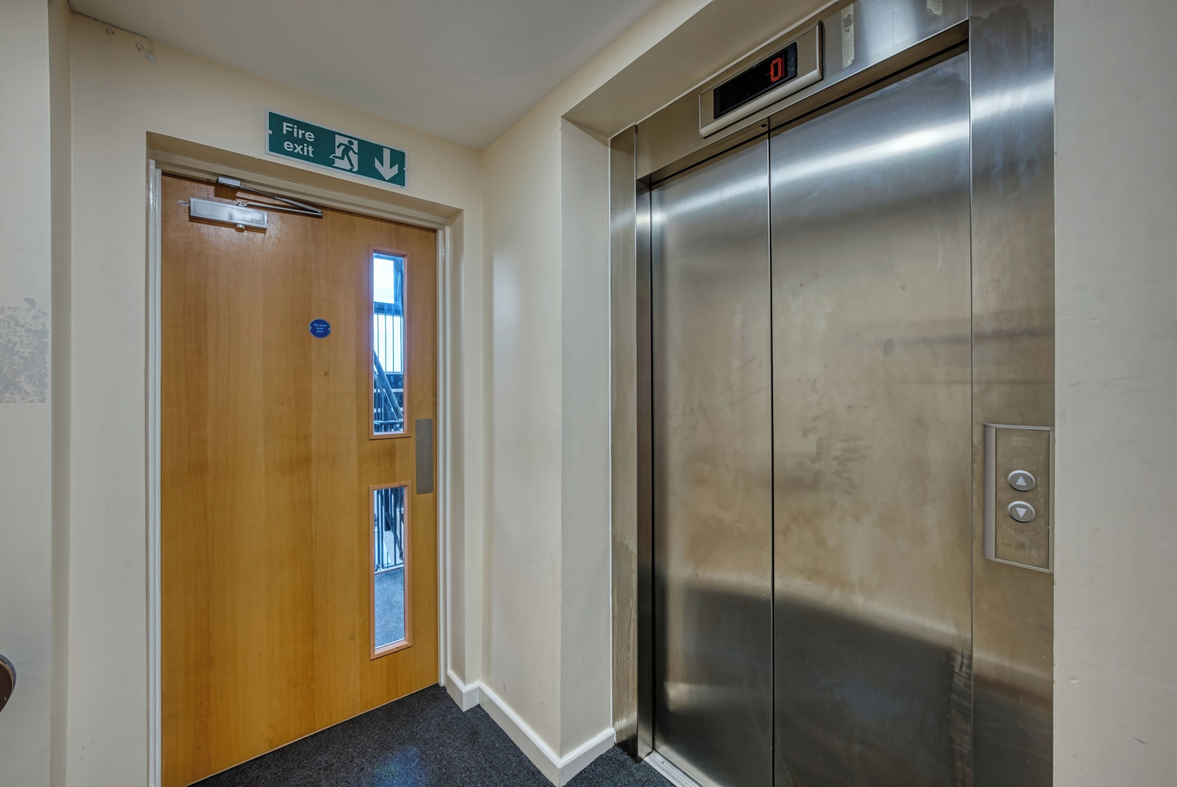 A hallway with a fire exit sign, a wooden door, and a stainless steel elevator.