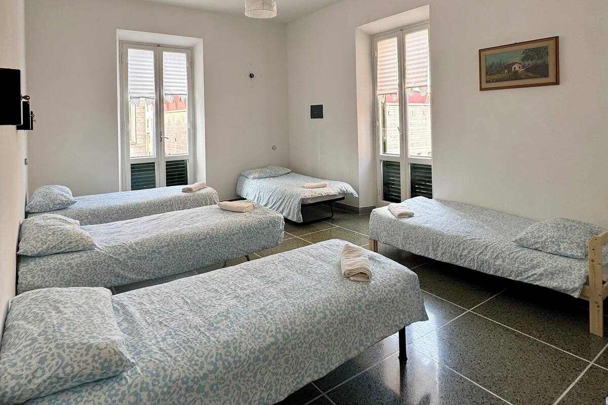 Une chambre de dortoir avec quatre lits simples, un luminaire au plafond et deux fenêtres équipées de stores.