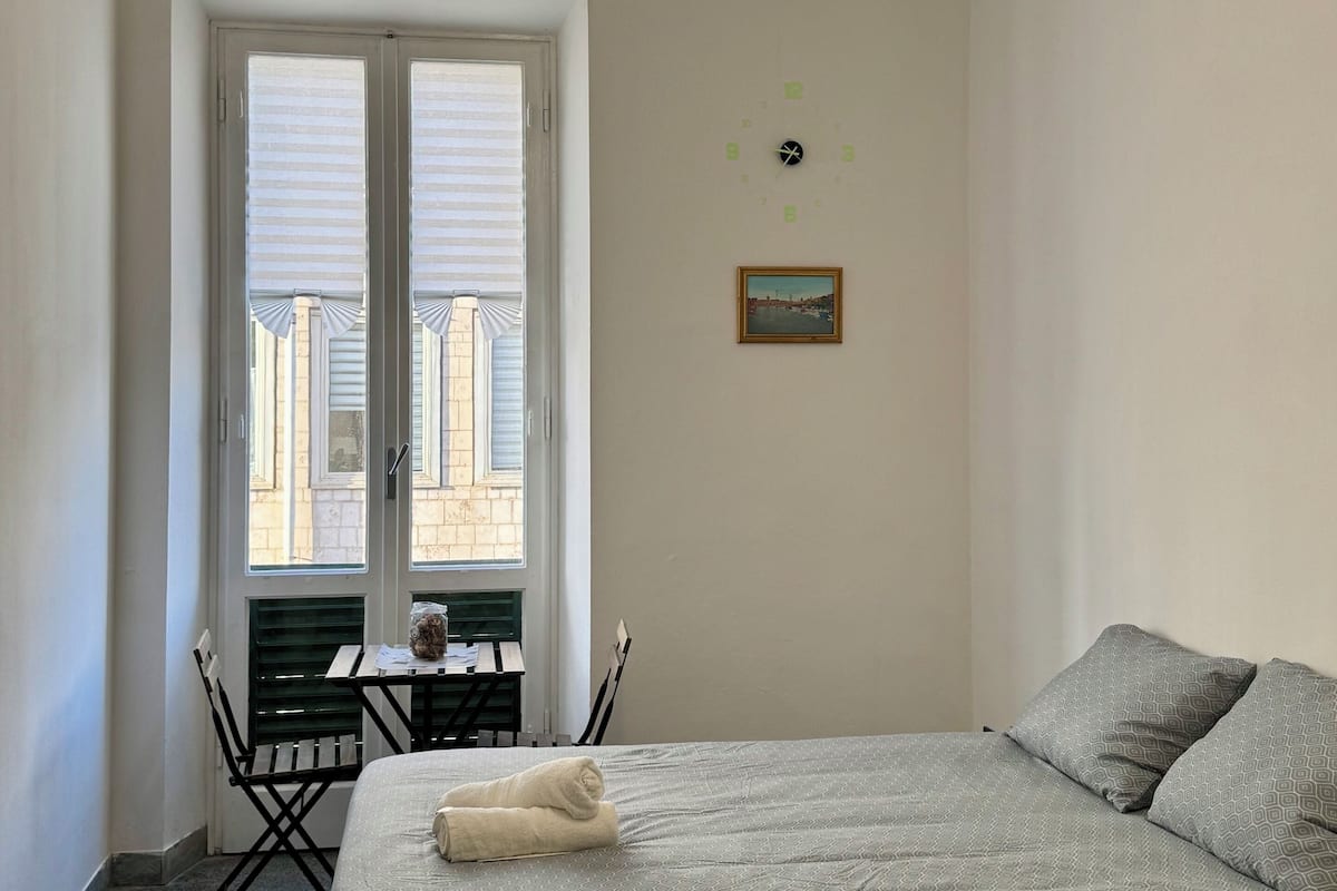 Une chambre à coucher avec un lit, une petite table et des chaises, une fenêtre avec des stores et un luminaire suspendu.