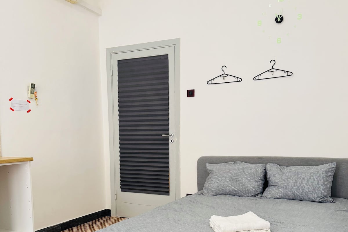 Une chambre à coucher avec un lit, une couette grise à motifs, un canapé gris avec des coussins à motifs, un sol à motifs, une armoire blanche et une porte avec un volet noir.