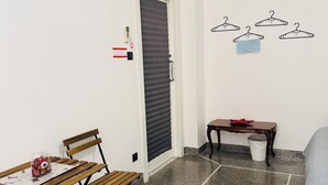 A small room with a wooden table, a bench, a wall-mounted air conditioner, a door with a striped roller blind, a small table with a red cloth, and a wall with four black hangers. - Affittacamere The Shuttle (Livorno)