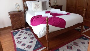 Un lit bien fait, avec une tête de lit en bois, du linge de lit blanc et des serviettes roses. On y trouve deux tables de chevet avec des lampes, une armoire en bois et des tapis à motifs sur le sol.