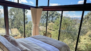Une chambre à coucher avec un lit, une tête de lit en bois et une vue sur une forêt, visible à travers de grandes fenêtres.