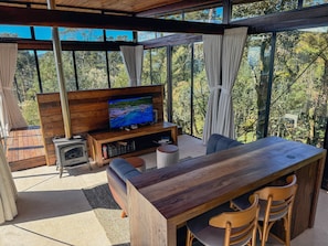 A modern outdoor living area with a wooden table and chairs, a television on a wooden stand, and a view of a forested landscape through large windows. - Serenus House (Urubici)