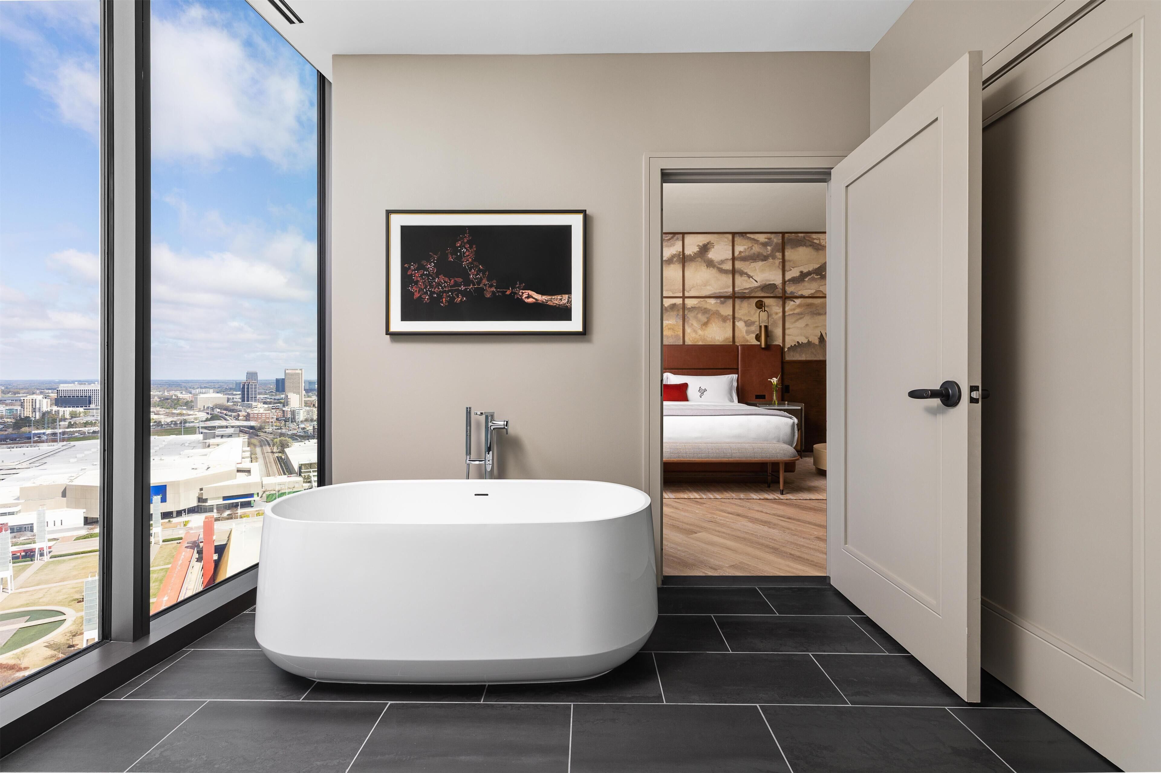 A modern bathroom with a large bathtub, a framed artwork, and a view of a cityscape.