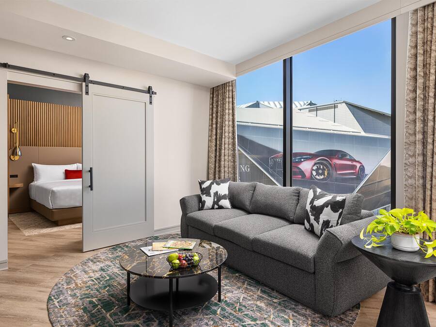 A modern living room with a grey sofa, a coffee table, and a view of a red sports car through large windows.