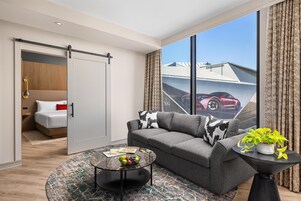 Una sala de estar moderna con un sofá gris, una mesa de centro y vistas a un coche deportivo rojo a través de amplios ventanales.