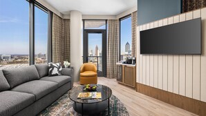 Un salon moderne avec un canapé, un fauteuil et une table basse, un téléviseur à écran plat fixé au mur, et une vue sur le paysage urbain à travers de grandes fenêtres.
