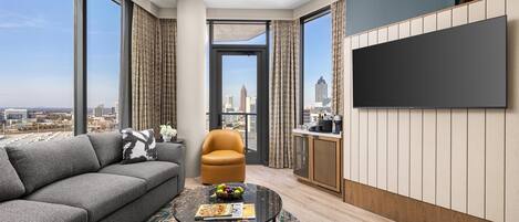 A modern living room with a sofa, armchair, and coffee table, a flat-screen TV mounted on the wall, and a view of the cityscape through large windows.