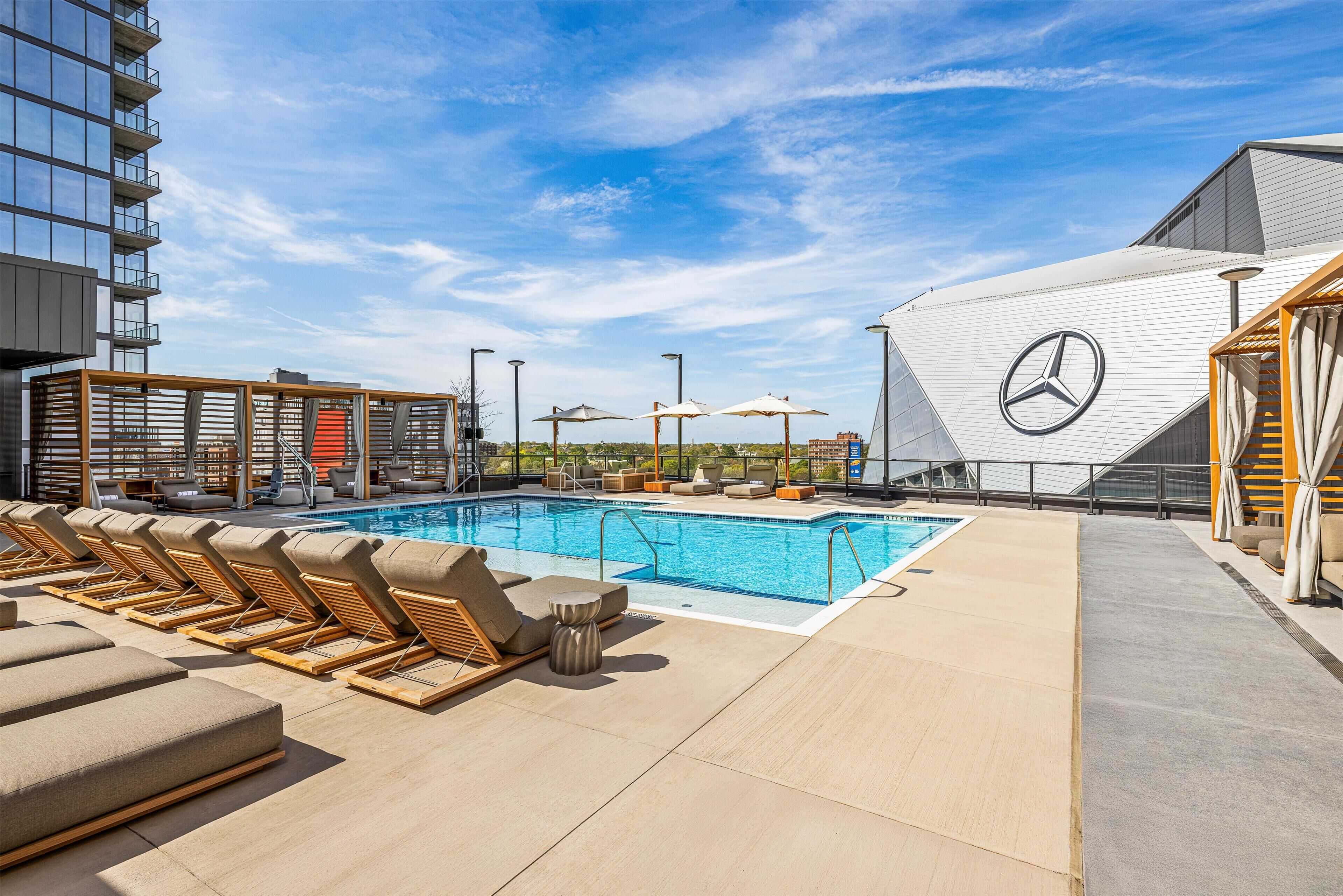 A rooftop pool area with lounge chairs and umbrellas, a Mercedes logo, and a modern building in the background.