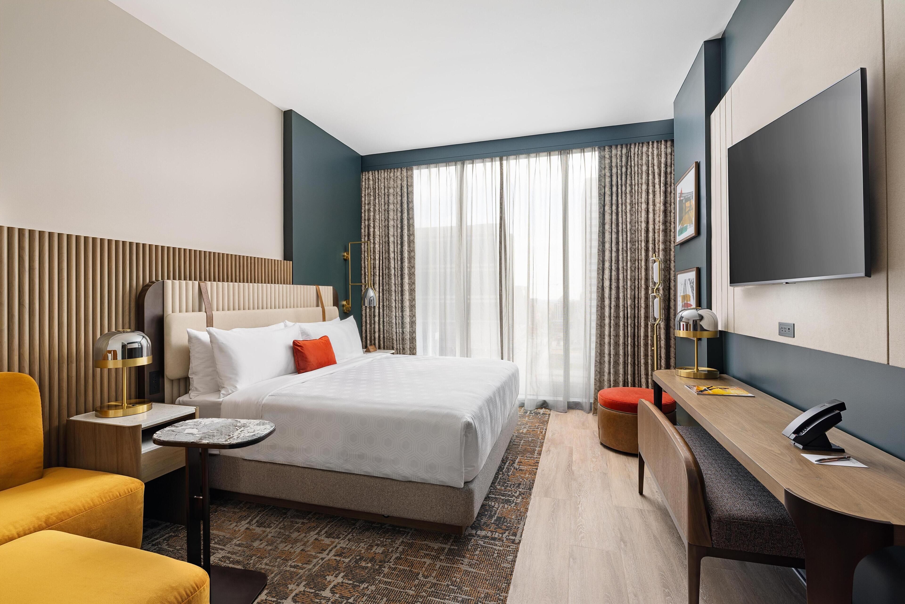 Une chambre d’hôtel avec un lit, un bureau, une chaise, une télévision et une fenêtre avec des rideaux.