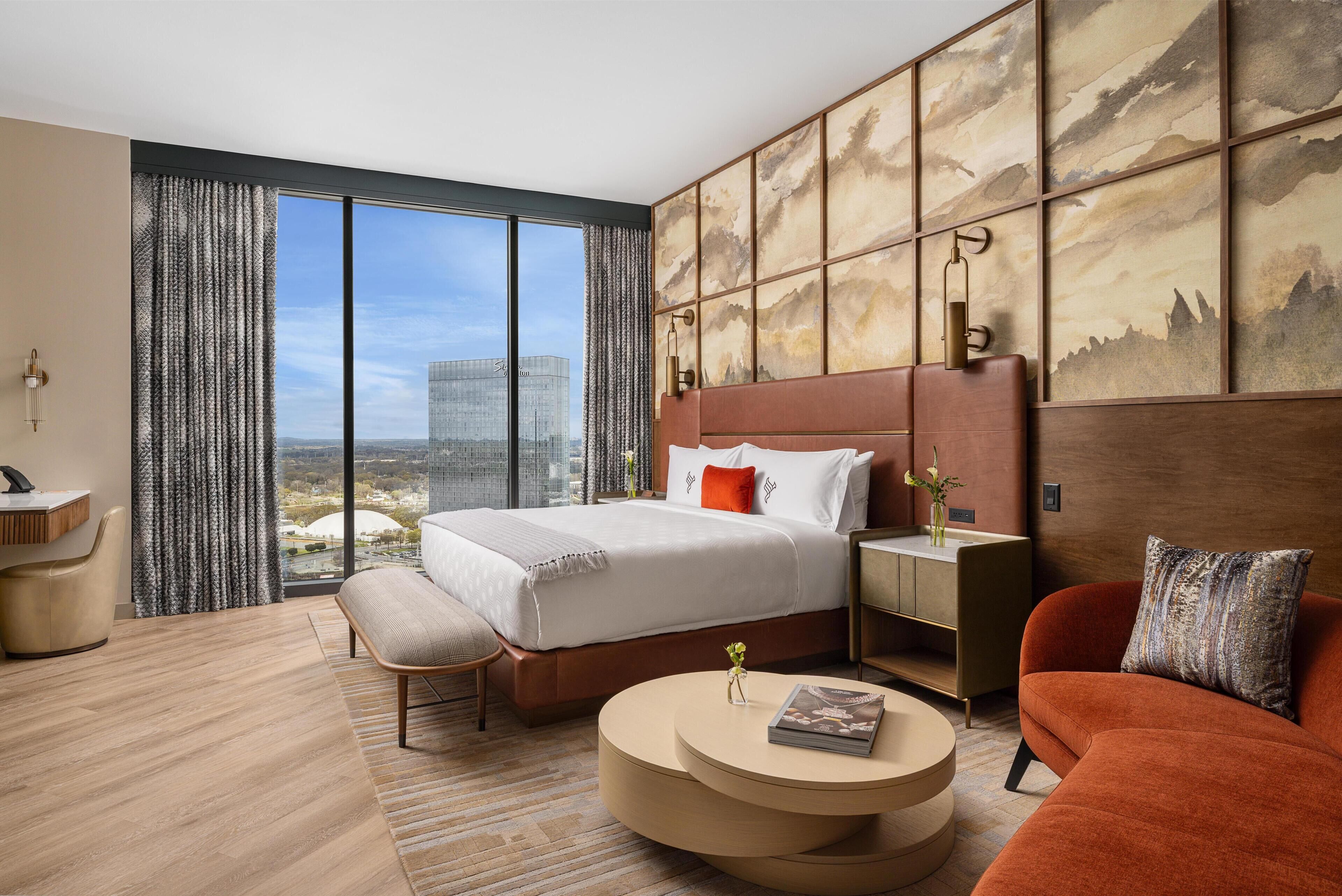 A modern hotel room with a large bed, a sofa, a round coffee table, and a view of the cityscape.
