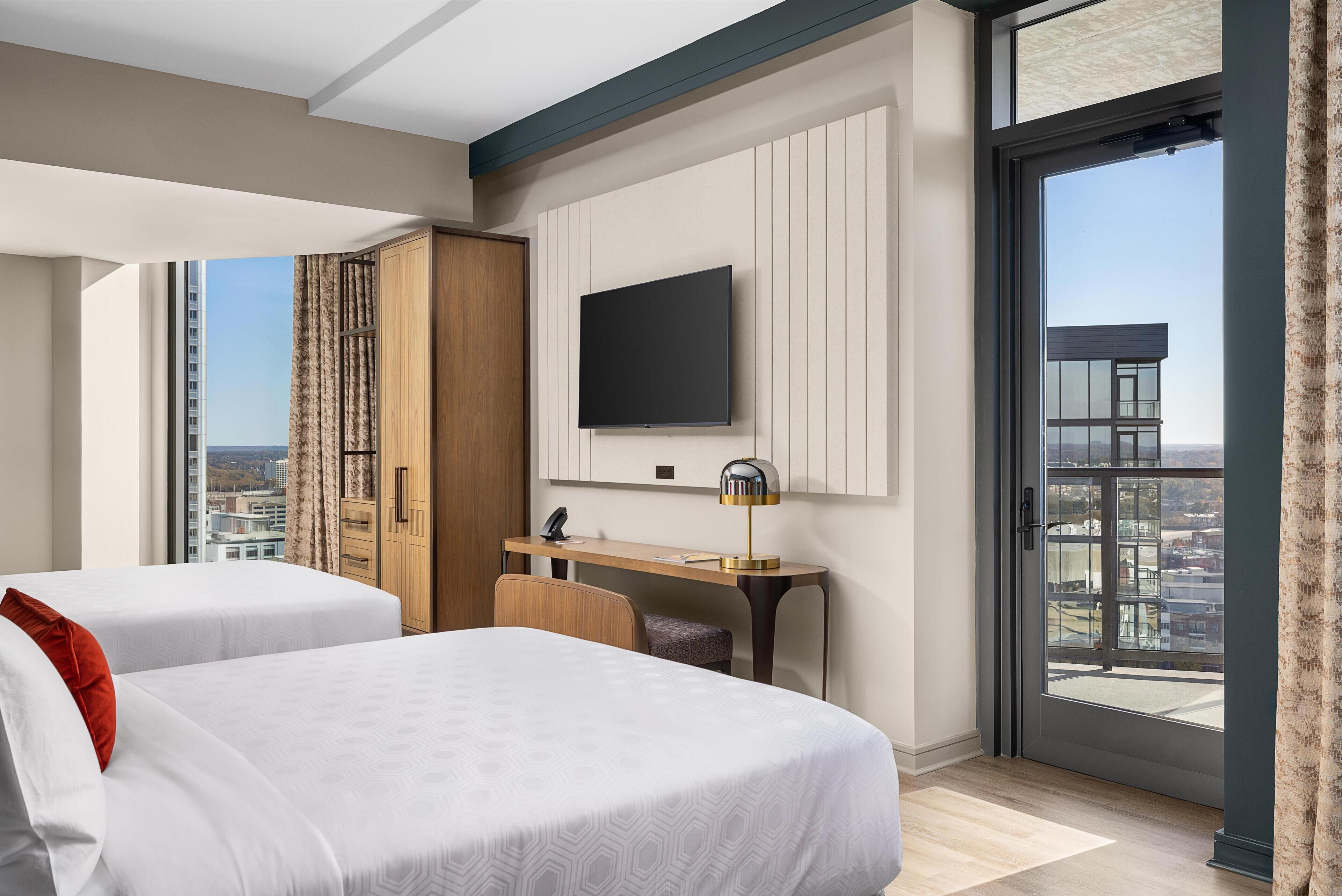A hotel room with two beds, a desk, a TV, and a large window with a city view.