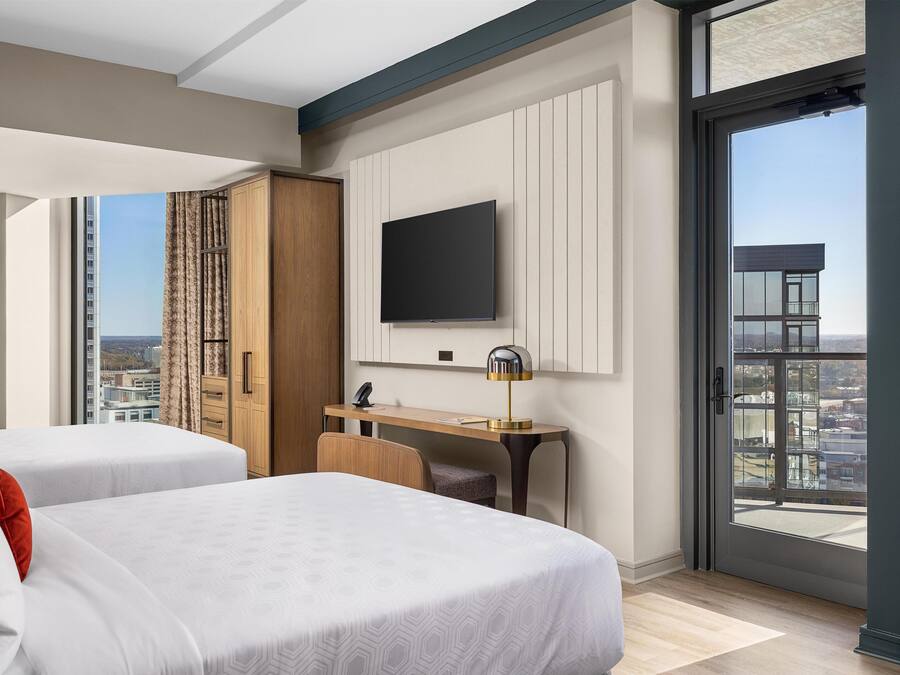 A hotel room with two beds, a desk, a TV, and a large window with a city view.