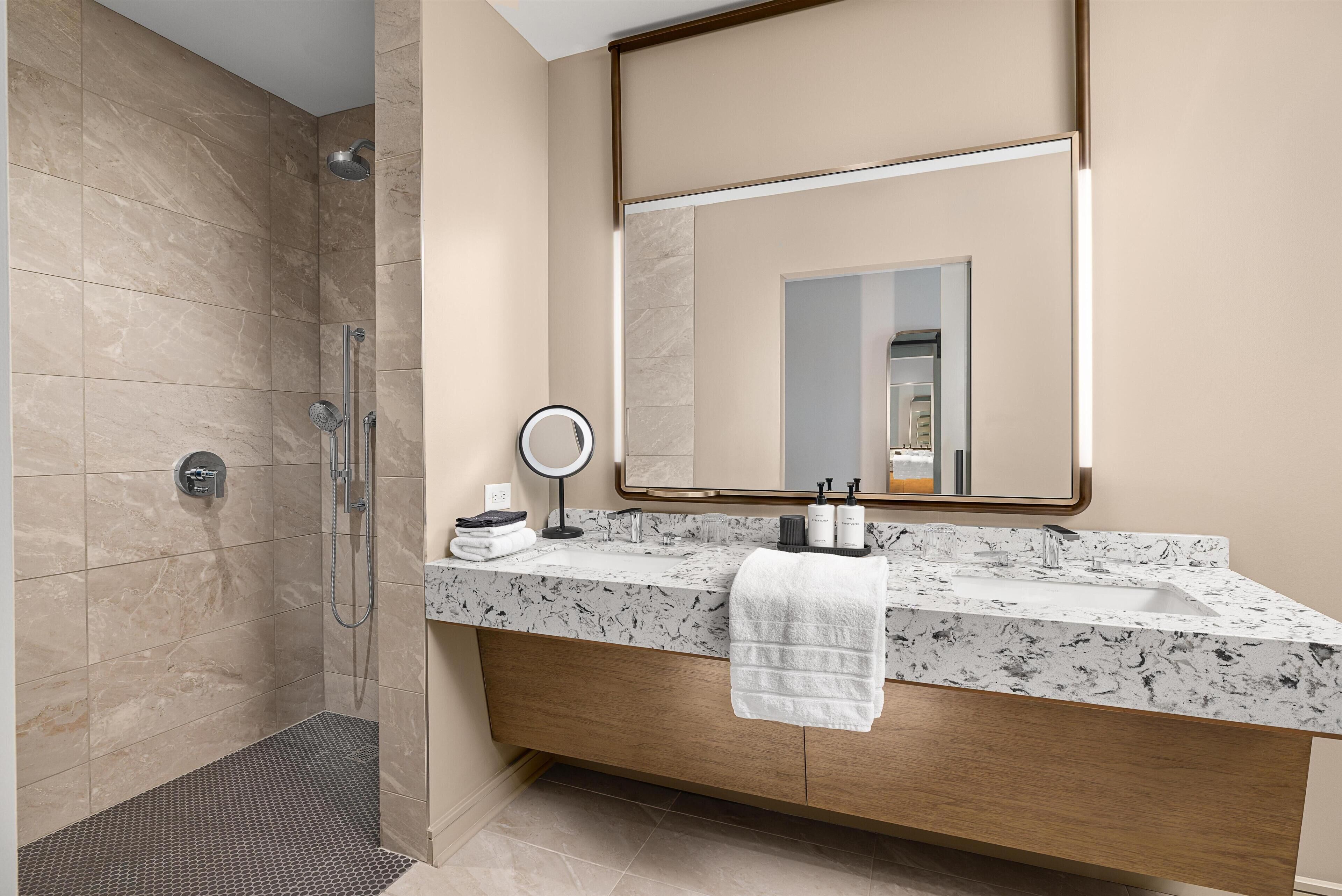 A modern bathroom with a shower, a vanity with a marble countertop, a large mirror, and a towel hanging on the wall.
