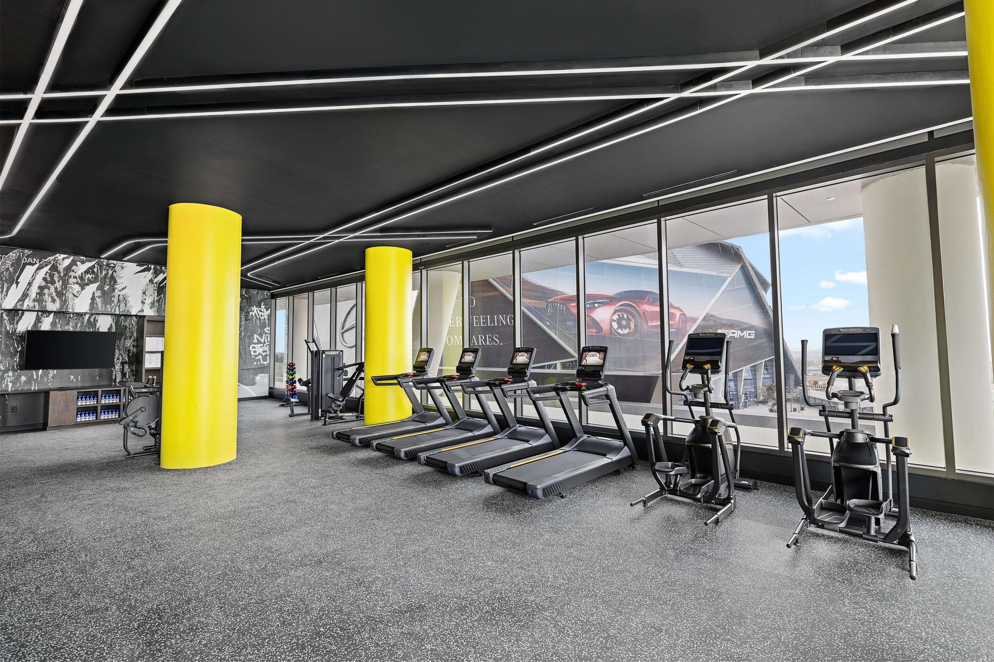 A gym with treadmills, elliptical machines, and a large window with a view of a red car and a building.