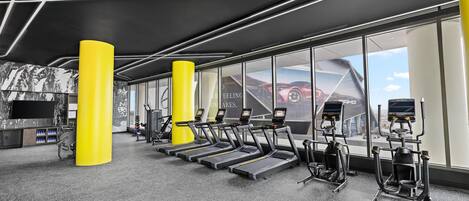 A gym with treadmills, elliptical machines, and a large window with a view of a red car and a building.
