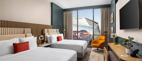 Habitación de hotel con dos camas, un escritorio, una silla, un televisor y un ventanal con vista a un coche rojo.