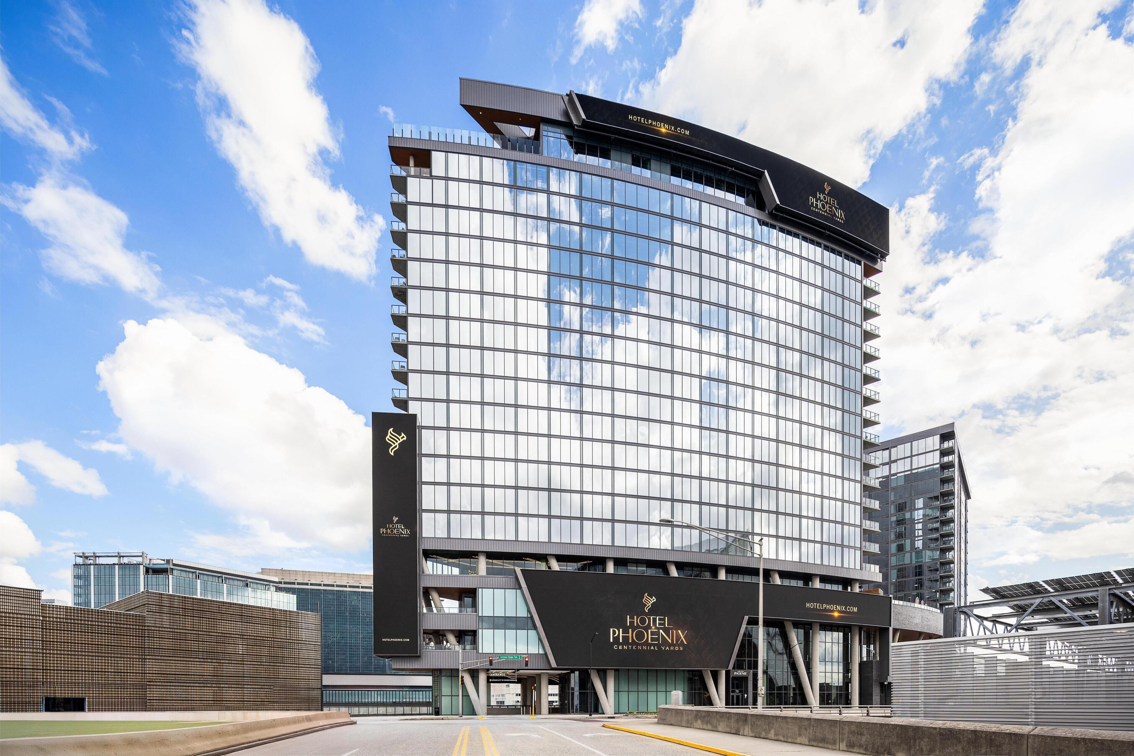 A modern hotel with a curved glass facade, a prominent sign reading 'Hotel Phoenix,' and a clear blue sky in the background.