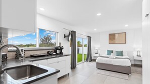 A modern kitchen with a built-in sink and a coffee machine, a bedroom with a bed and a nightstand, and a large window with curtains. - Apt W/patio, Balcony & Mini-golf (Deerfield Beach)