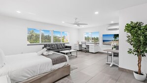 A modern hotel room with a bed, a desk, a chair, a sofa, and a large window with a view of the beach.