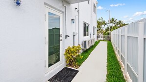 A white building with a narrow pathway, air conditioning units, and a white fence.