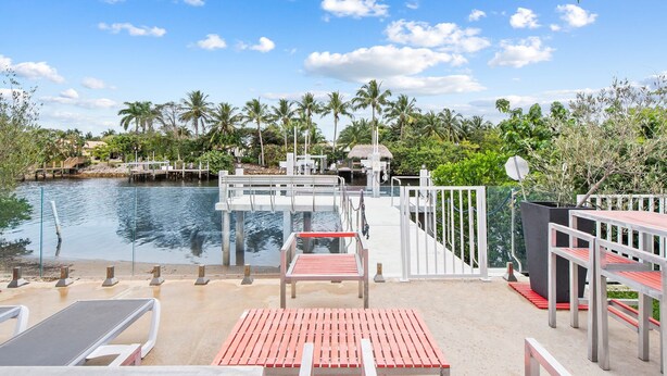 A waterfront patio with wooden seating, a pool, and palm trees in the background.