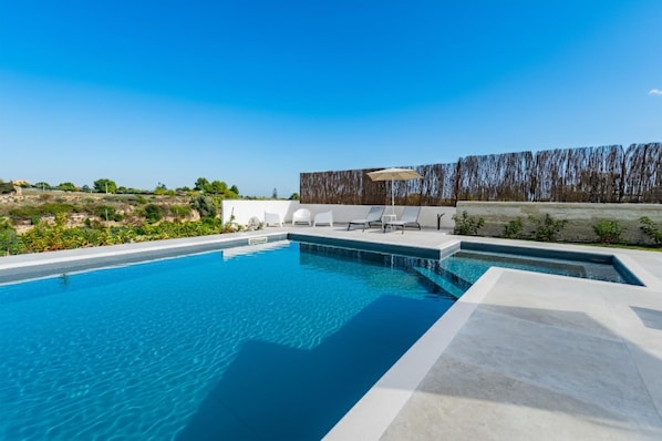 Una piscina con agua cristalina azul, rodeada de sillas reclinables blancas y una valla de madera.