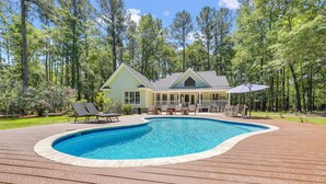 Une maison avec une piscine, entourée d’une terrasse en bois, d’un patio couvert et bordée par une forêt.