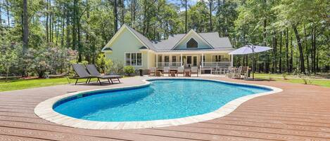 A house with a swimming pool surrounded by a wooden deck, a covered patio, and a forested backdrop.