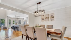 Un coin repas avec une table en bois, six chaises et un lustre. Des tableaux encadrés ornent le mur, et on aperçoit une cuisine au fond.