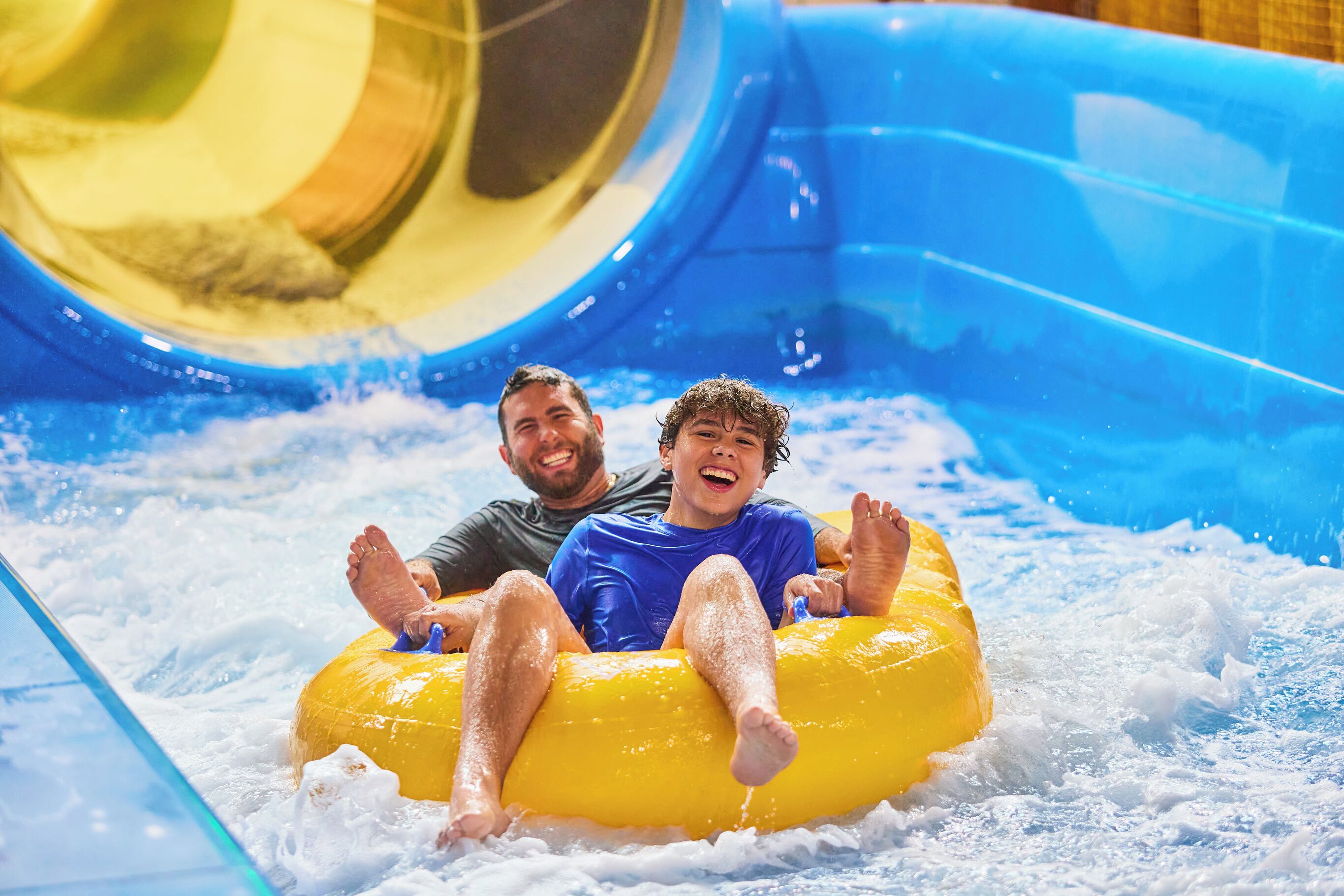 Zwei Personen auf einem gelben Luftschlauch in einem Wasserspielbereich mit einer blauen Rutsche.