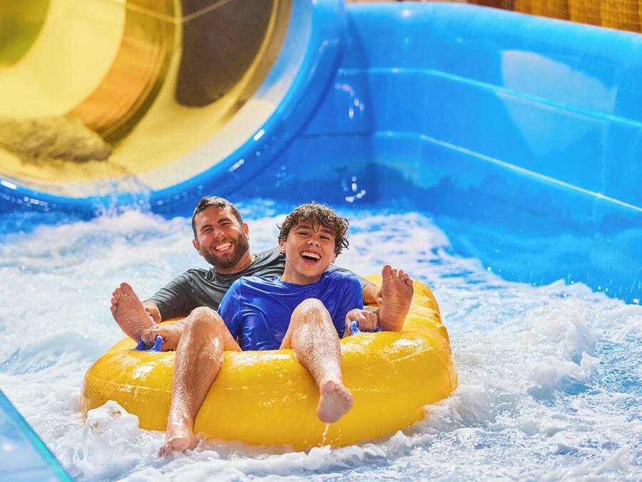 Duas pessoas em um boião amarelo em uma área de recreação aquática com um escorregador azul.