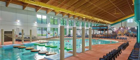 An indoor swimming pool with multiple lanes, a diving board, and a seating area with chairs.