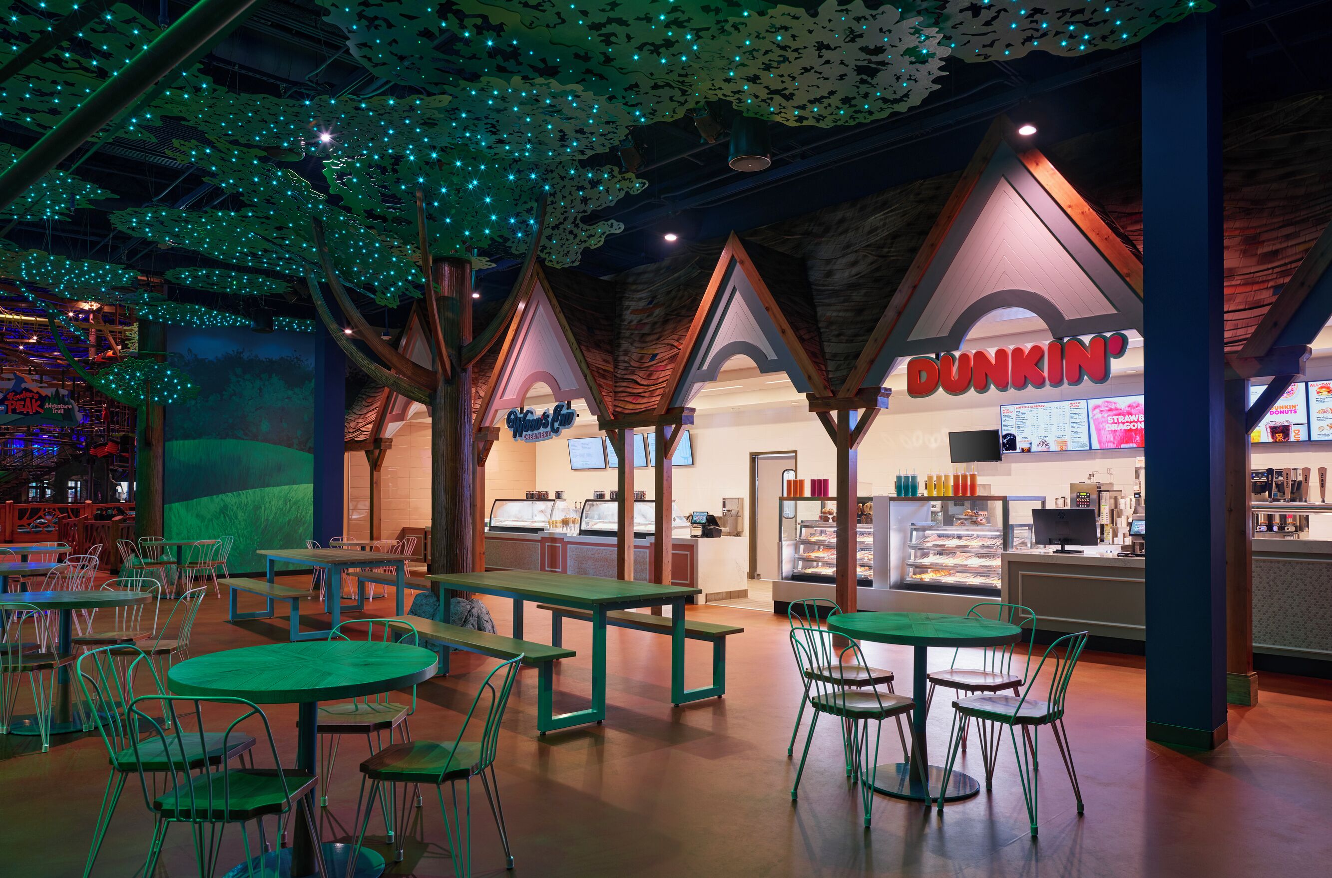 An indoor dining area with green tables and chairs, a Dunkin' Donuts counter, and a ceiling with hanging lights.