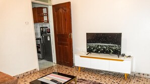 Ein Wohnzimmer mit einem Fernseher auf einem weißen Schrank, einem Couchtisch mit einer Fernbedienung und einer Tür zu einer Küche.