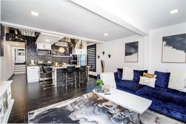 A modern kitchen with a marble backsplash, a dining area with a white table and blue sofa, and a living room with dark flooring and a large rug. - Heated Pool, Spa (Clearwater)