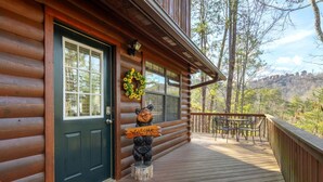 A log cabin with a dark front door, a welcome sign, a bear statue, a wreath, and a deck with outdoor furniture. - Bear Hideaway (Sevierville)
