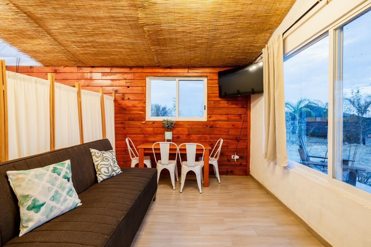 Een gezellige kamer met een houten plafond, een bruine bank met gestreept kussens, een eettafel met witte stoelen en een groot raam met uitzicht op de omgeving.